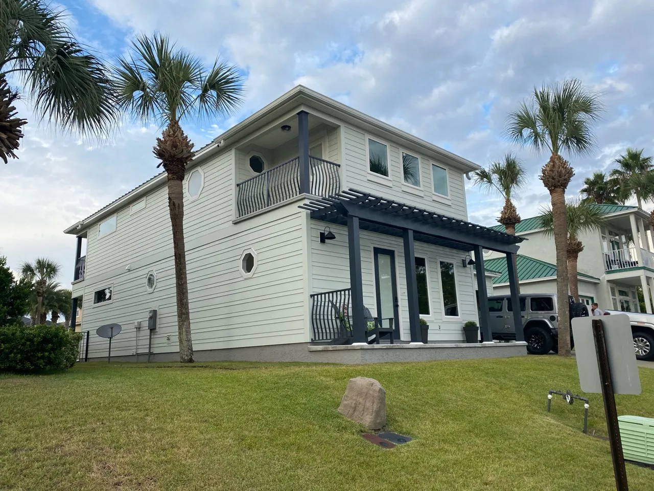 A completed two-story home in Panama City Beach with pale siding, a dark pergola over the front porch, and palms in the foreground.